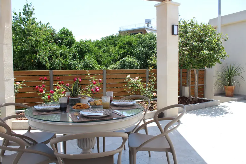 The round table under the pergola laid for breakfast, red roses in the bed behind it