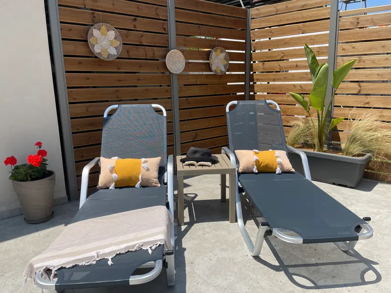 Two grey sun beds with yellow cushions against a wooden slatted fence, a red geranium in a pot beside them